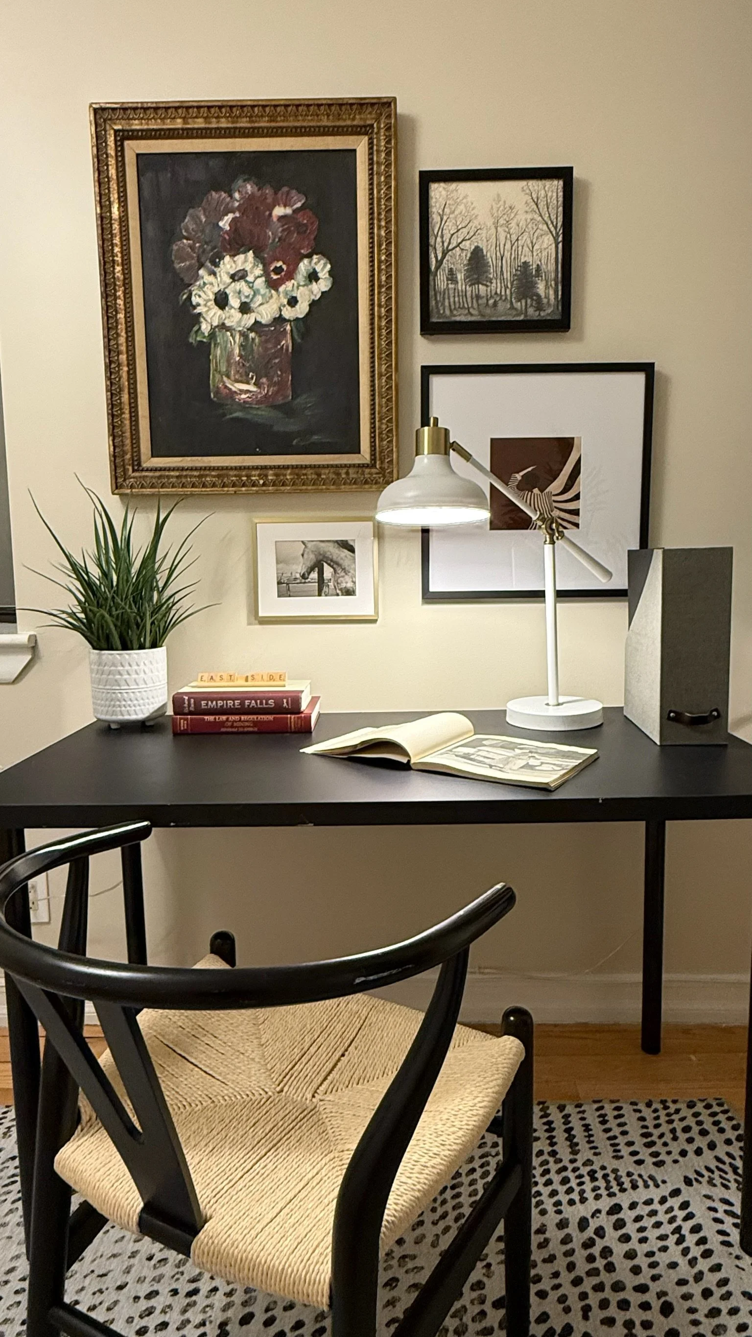 A black table with a black chair that has a woven beige seat. On the table, there are three stacked books, an open book, a potted plant, a desk lamp, and a gray speaker. Behind the table, a wall features five framed artworks, including a large floral painting and various monochrome and abstract art pieces, with part of a polka-dotted rug visible in the foreground.