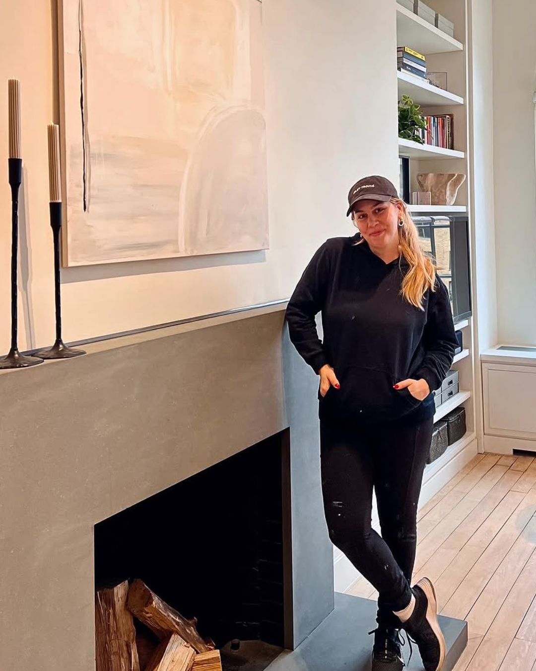 A woman standing casually by a modern fireplace with neatly stacked firewood inside, in a contemporary living room. She's wearing a black hoodie, black pants, and a black baseball cap, with her hands in her pockets.
