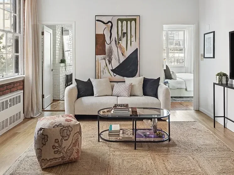 Living room with beige sofa, dark pillows, abstract art on the wall, rectangular glass coffee table, upholstered ottoman, and wooden flooring.