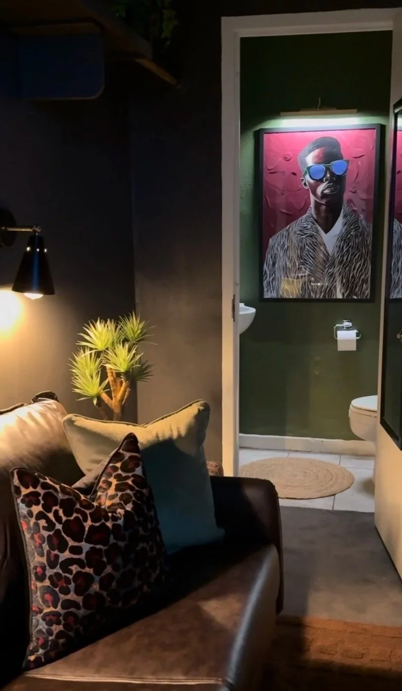 Interior view of a living room with a dark-colored wall, a leather sofa with animal print and plain cushions, a small potted pine tree, a wall-mounted lamp, and a door leading to a bathroom. The bathroom has a portrait of a person wearing sunglasses and a fur-patterned coat on the wall.