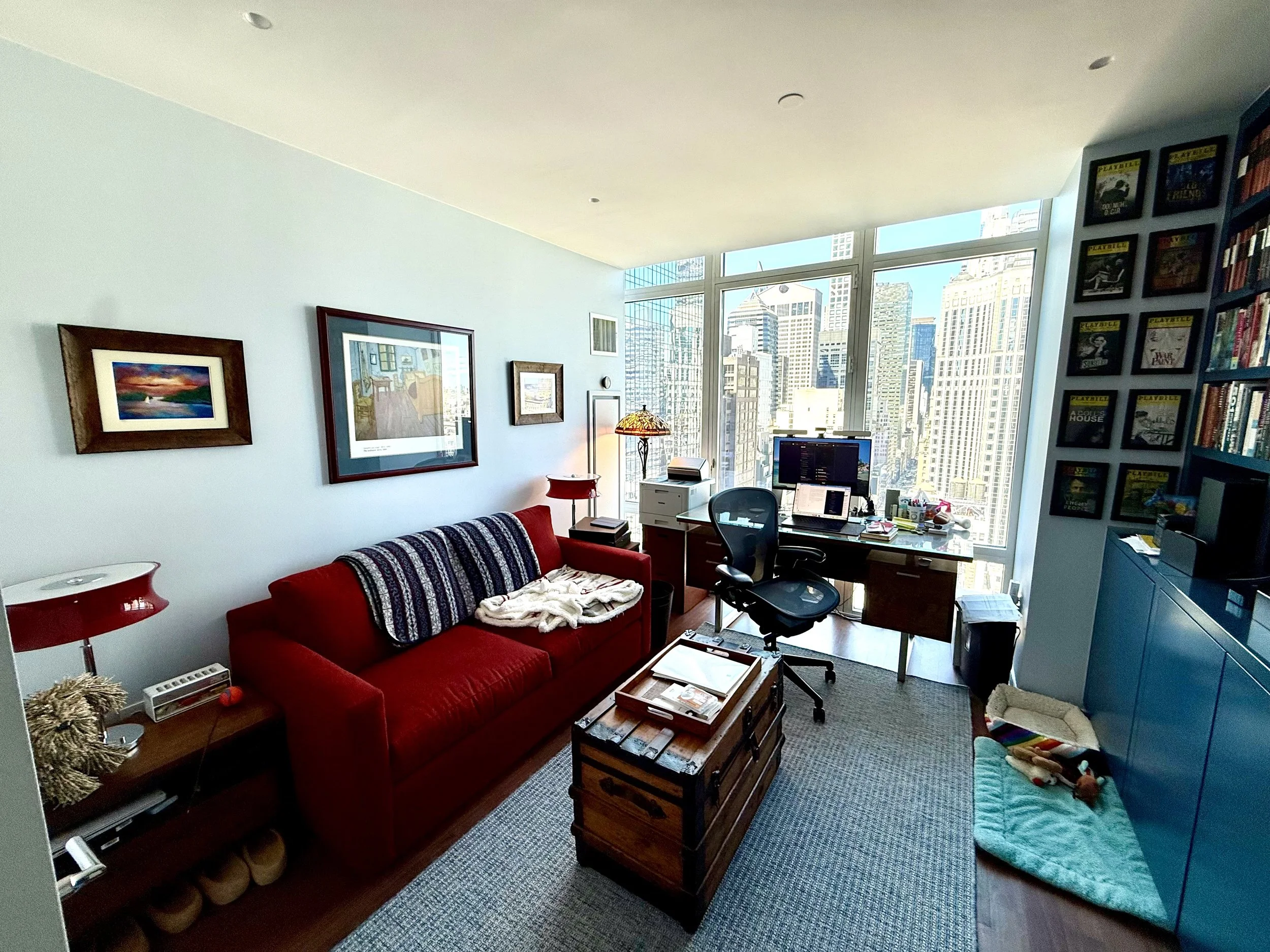 A cozy urban apartment living room with large windows showing city skyscrapers, a red sofa with a striped blanket, framed artwork on the pastel blue wall, a blue bookshelf filled with books, a desk with a computer and office supplies, a colorful knot rug, and a teal pet bed with a dog toy.