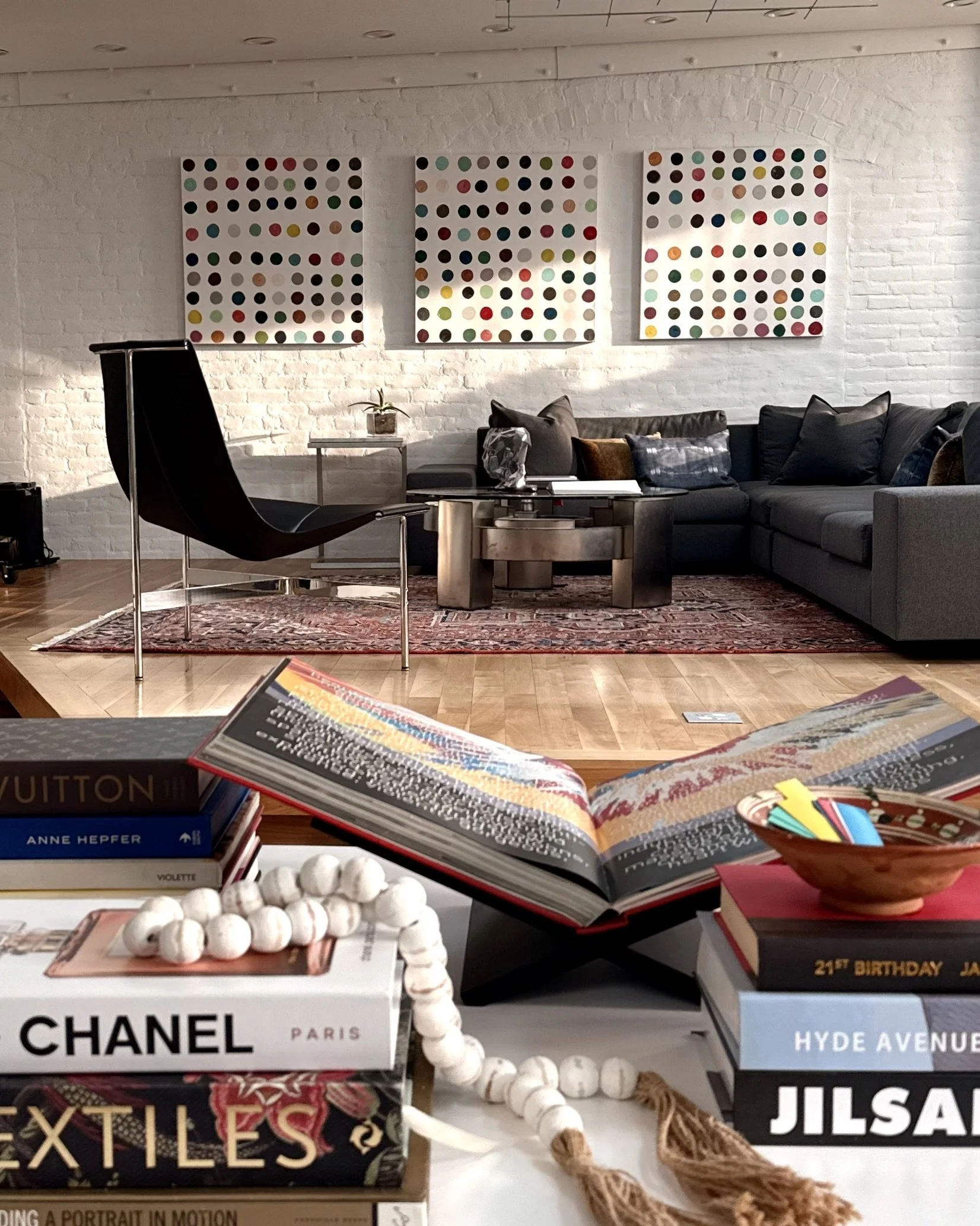 Display of fashion and lifestyle books, including brands like Chanel, Jil Sander, and Jilsa, with decorative elements such as a string of white beads and a rug, in a modern interior setting with white brick walls and a sofa.