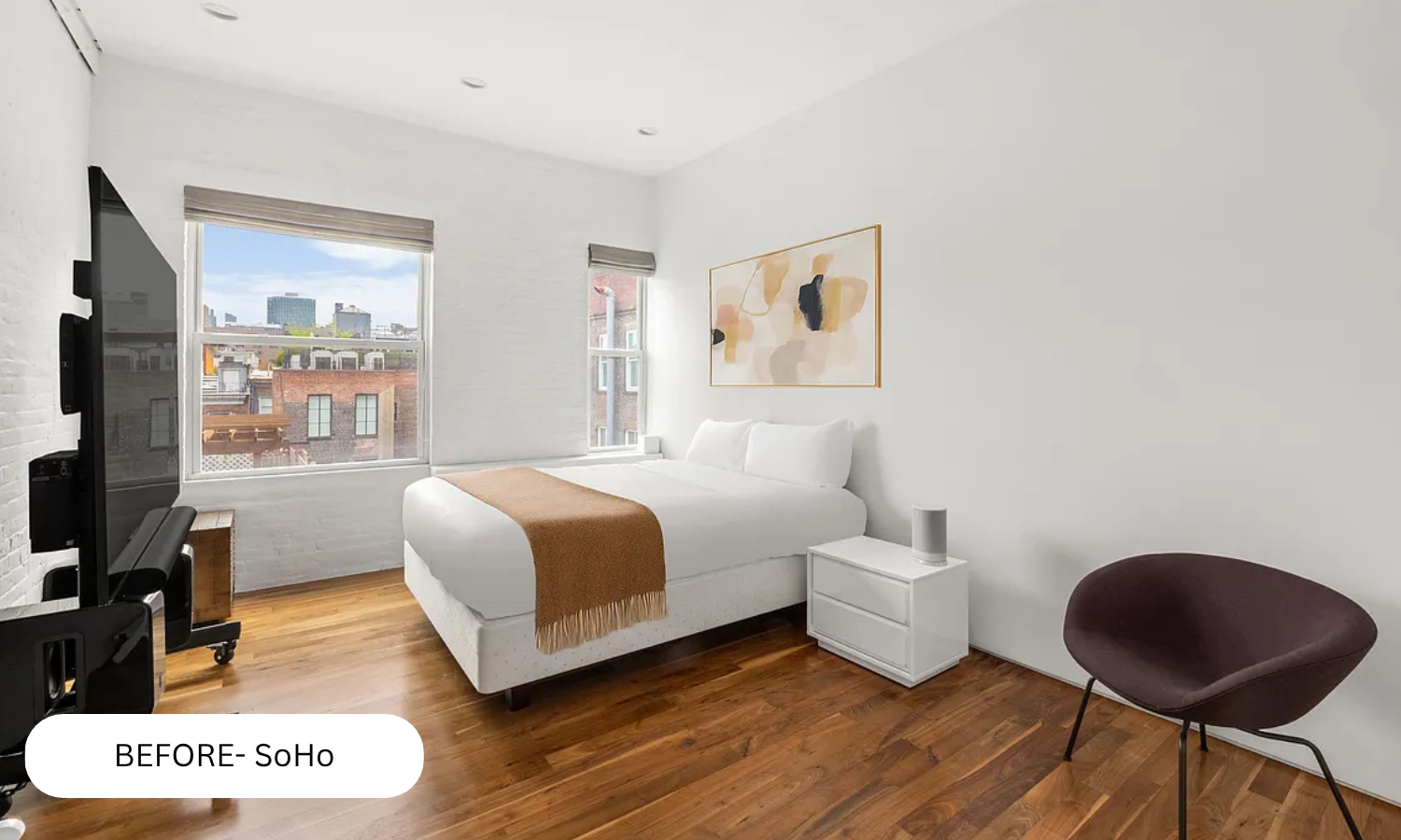 Minimalist bedroom with white walls, hardwood floor, white bed with beige throw, abstract art, brown chair, white nightstand, TV, and window with city view.