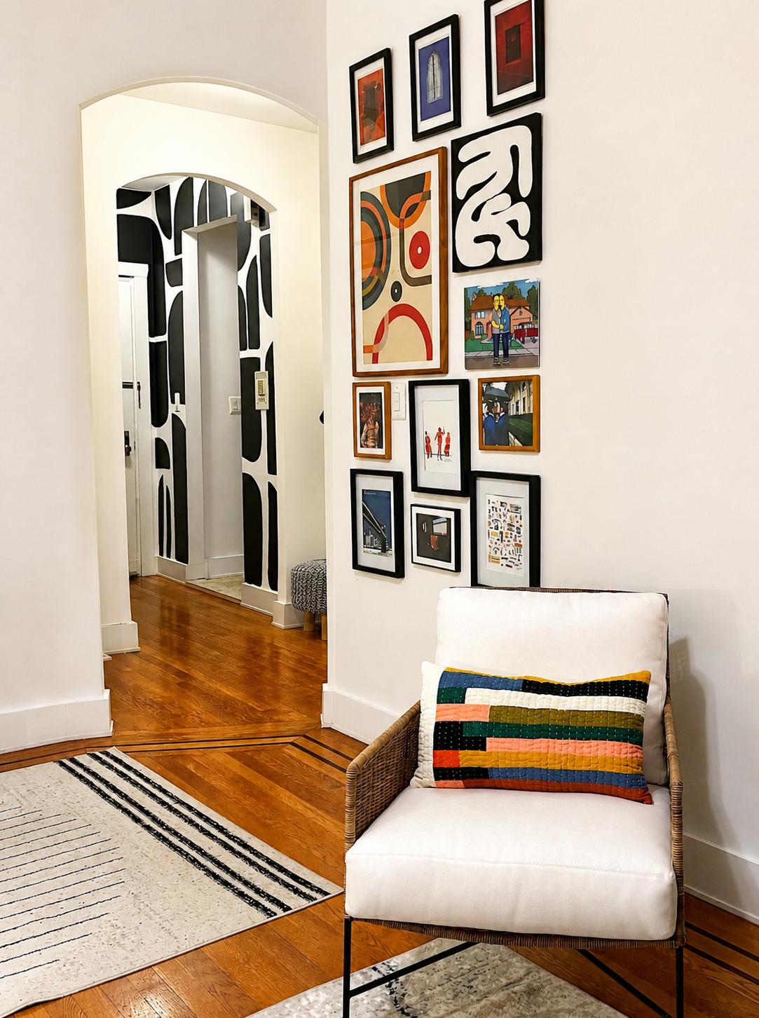 A gallery wall with various framed artwork and photos in a well-lit room, a white chair with a colorful, striped pillow, a black-and-white striped rug, and hardwood floors.