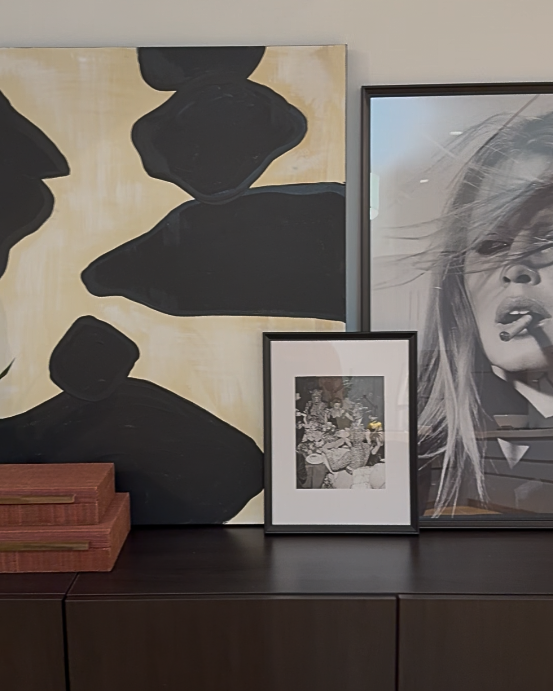 Decorative wall art, including a large abstract painting with black and beige shapes, a black-and-white photograph of a woman smoking, and a smaller framed black-and-white photograph of people at a banquet, displayed on a dark wooden sideboard.