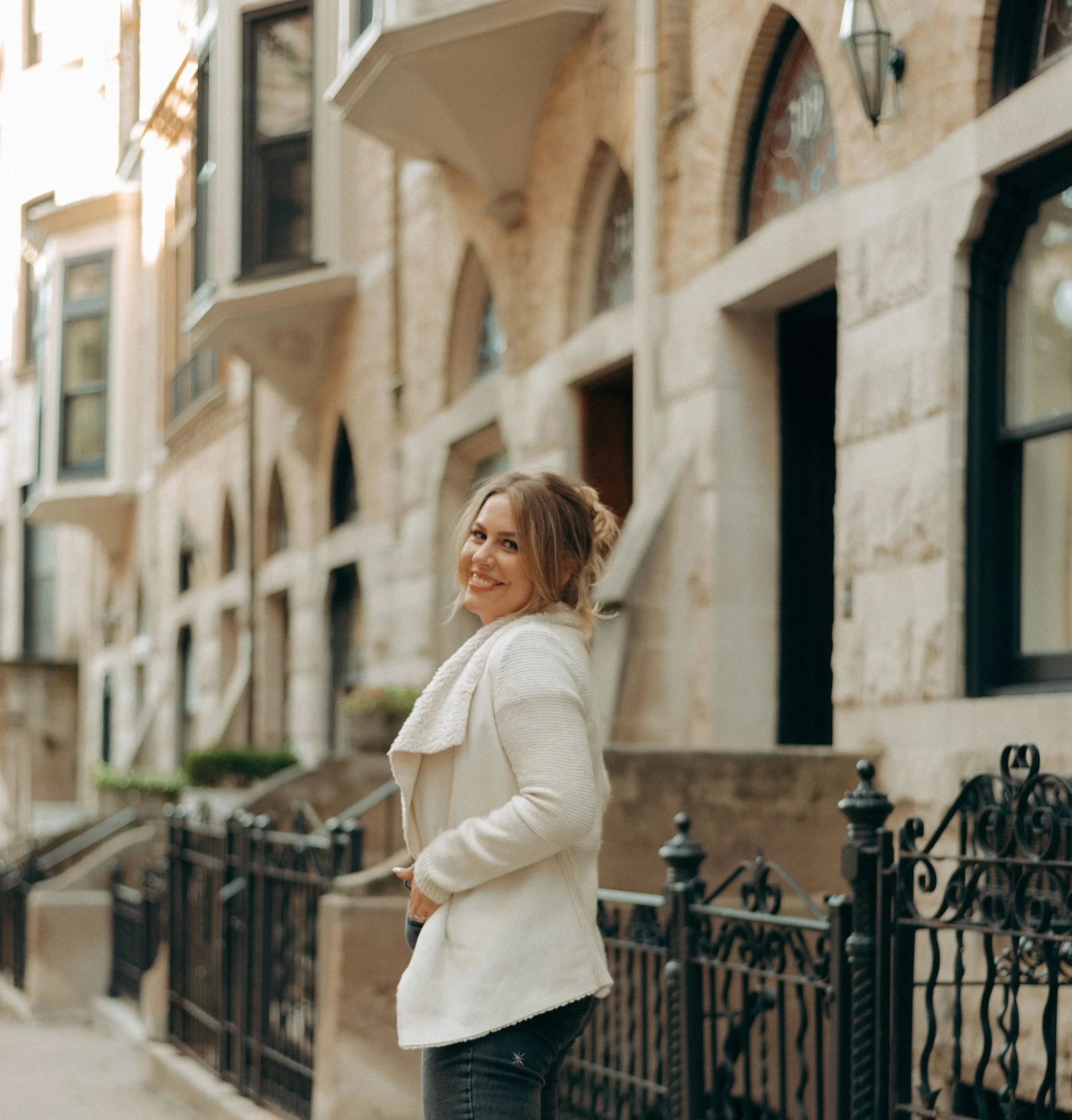A woman with light brown hair, wearing a cream-colored sweater and dark jeans, smiling and standing outdoors in front of a decorative wrought iron fence and a brick building with arched windows in New York City.