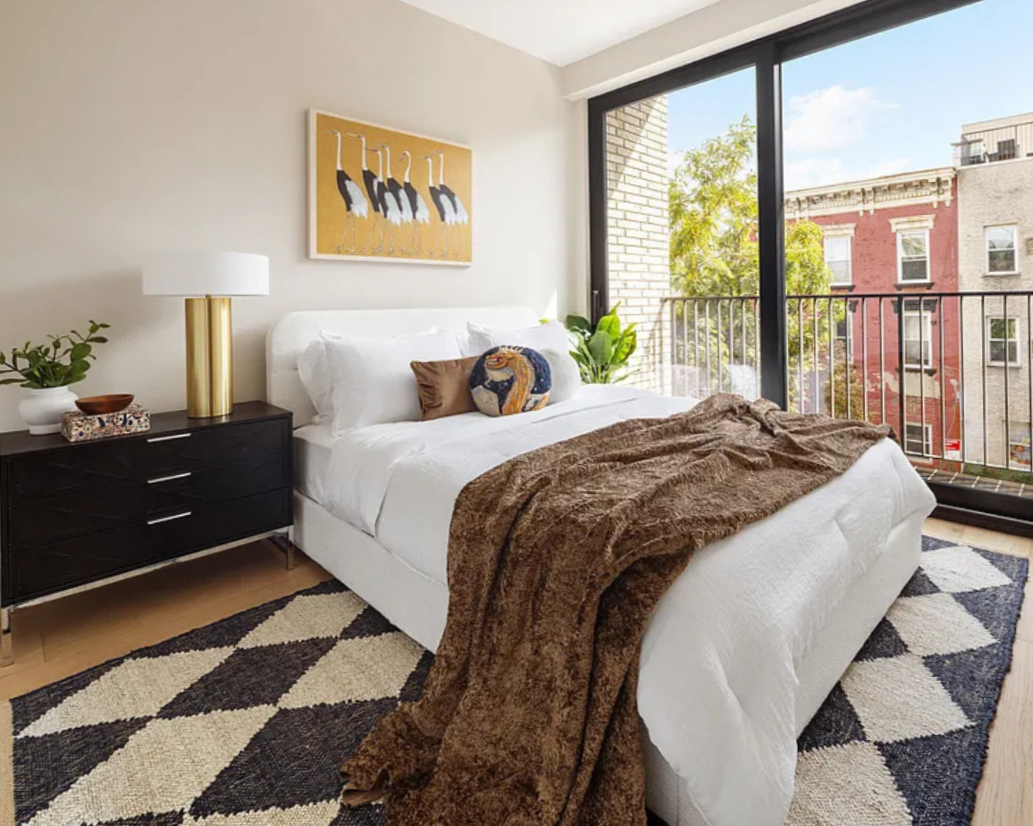 A bright bedroom with a white bed, brown blanket, decorative pillows, black nightstand with a gold lamp, plant, and art on the wall. Large glass sliding doors open to a balcony with city buildings and trees outside.