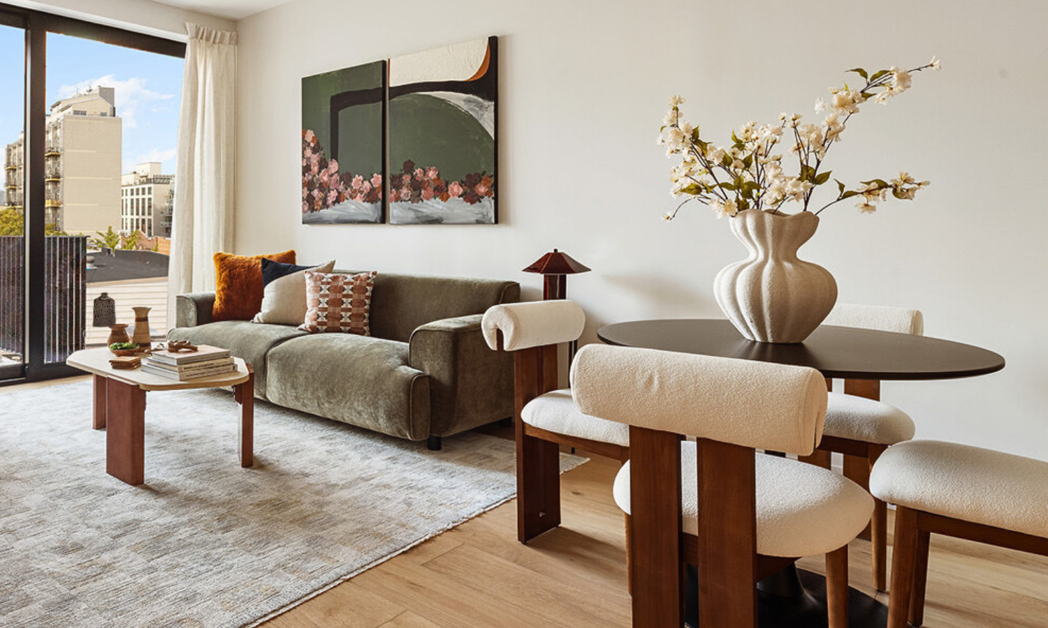 Living room with a sofa, a round dining table with a vase of flowers, and a balcony door with city buildings outside.