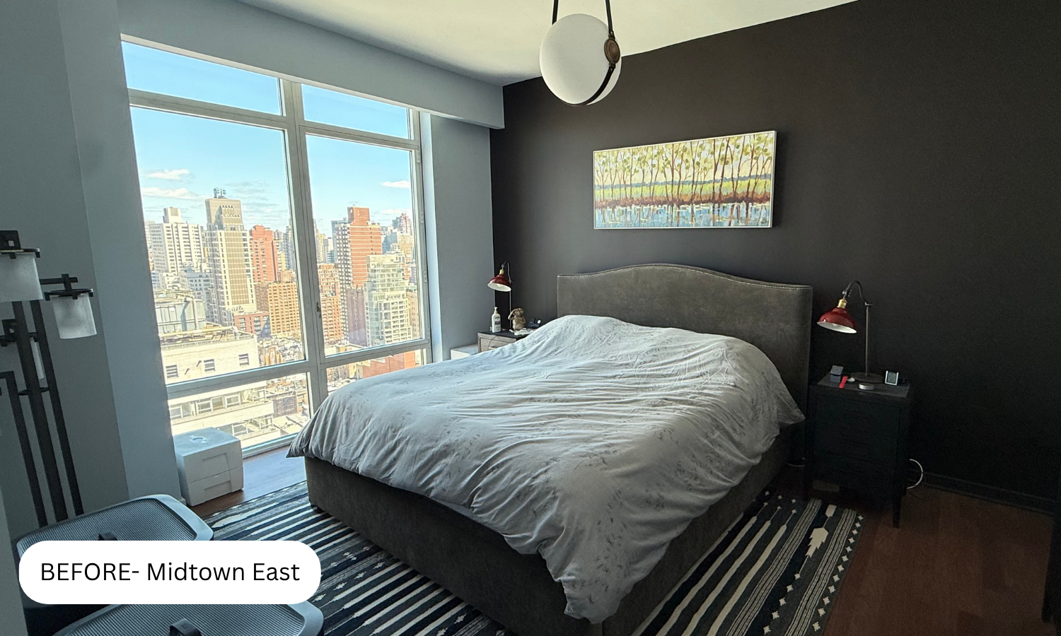 A bedroom with a large window overlooking a cityscape of tall buildings. The room has a dark accent wall behind the bed, a painting above the bed, bedside lamps, and a black nightstand. The bed has a grey headboard and light-colored bedding.