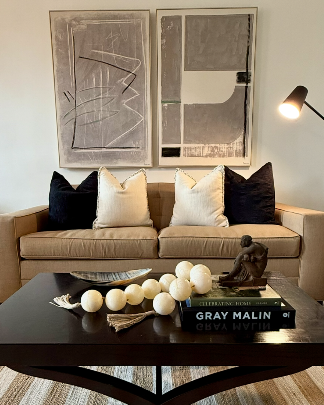 A beige sofa with two white and two black pillows in front of two framed abstract art pieces on a white wall. A black coffee table with decorative objects, including white spherical beads, a black sculpture, a tray, and books, is in front. A black floor lamp is on the right side.