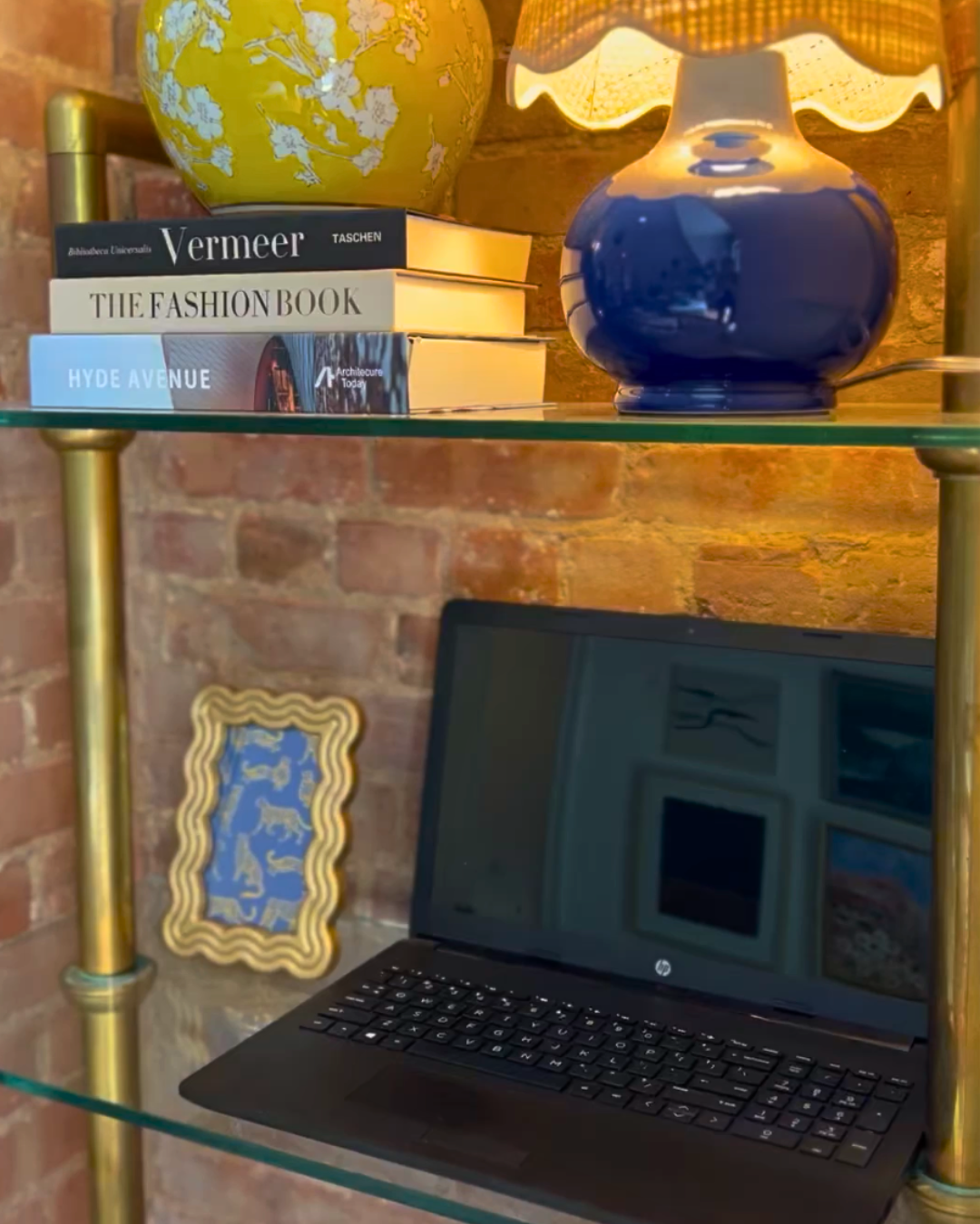 A glass shelf with a yellow and white floral vase, four stacked books titled 'Vermeer,' 'The Fashion Book,' 'Hyde Avenue,' and 'Archinect Today,' a blue ceramic lamp with a yellow lampshade, a brick wall in the background, a black laptop, and a decorative golden frame with a blue and gold design leaning against the wall.