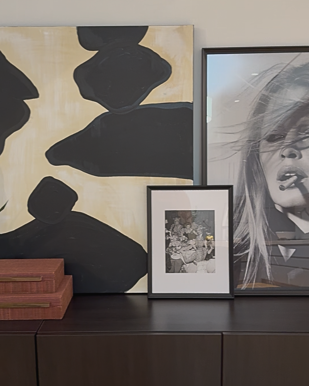 A gallery wall with three framed pictures, a large black-and-white photo of a woman smoking, an abstract painting with black shapes on a beige background, and a small black-and-white photo featuring people and flowers, all arranged on a dark wooden surface.