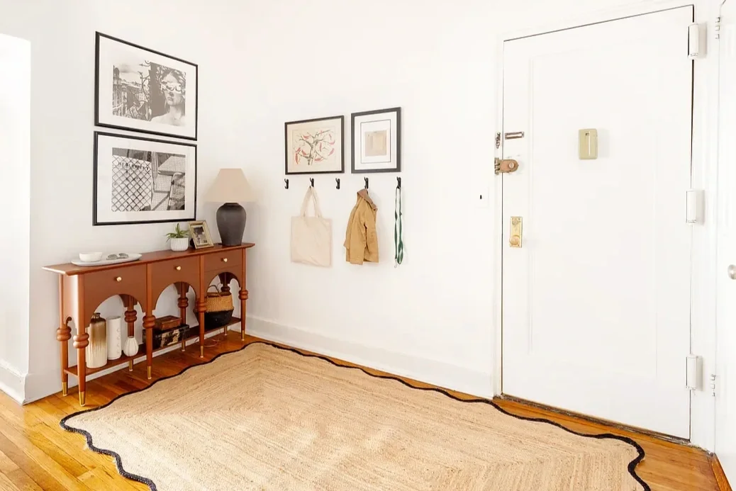 Entryway with a wooden console table, decorative vases, and a lamp, guide hooks with purses and a coat, framed wall art, and a beige rug on wooden flooring.