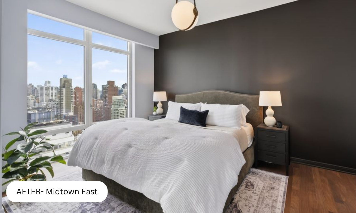Modern bedroom with large windows showing city skyline, dark accent wall, white bedding, two nightstands with lamps, and hardwood floors.