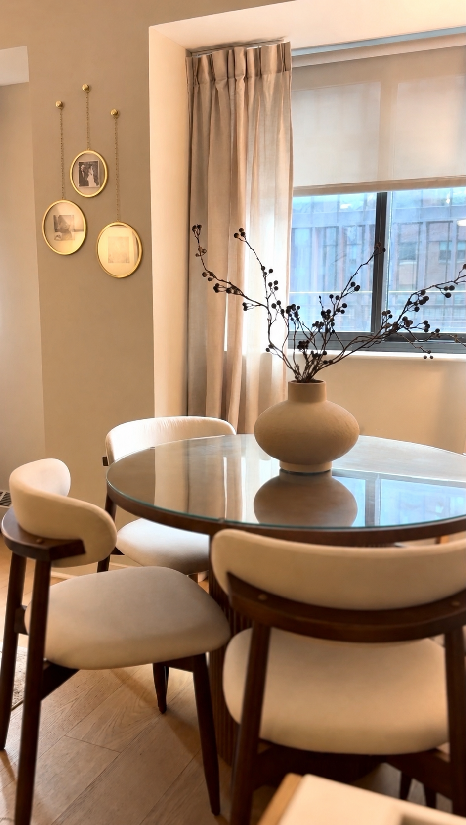 A round glass dining table with four mid-century modern chairs, a beige vase with dried branches, beige curtains, a window, and a wall display of three hanging oval picture frames.