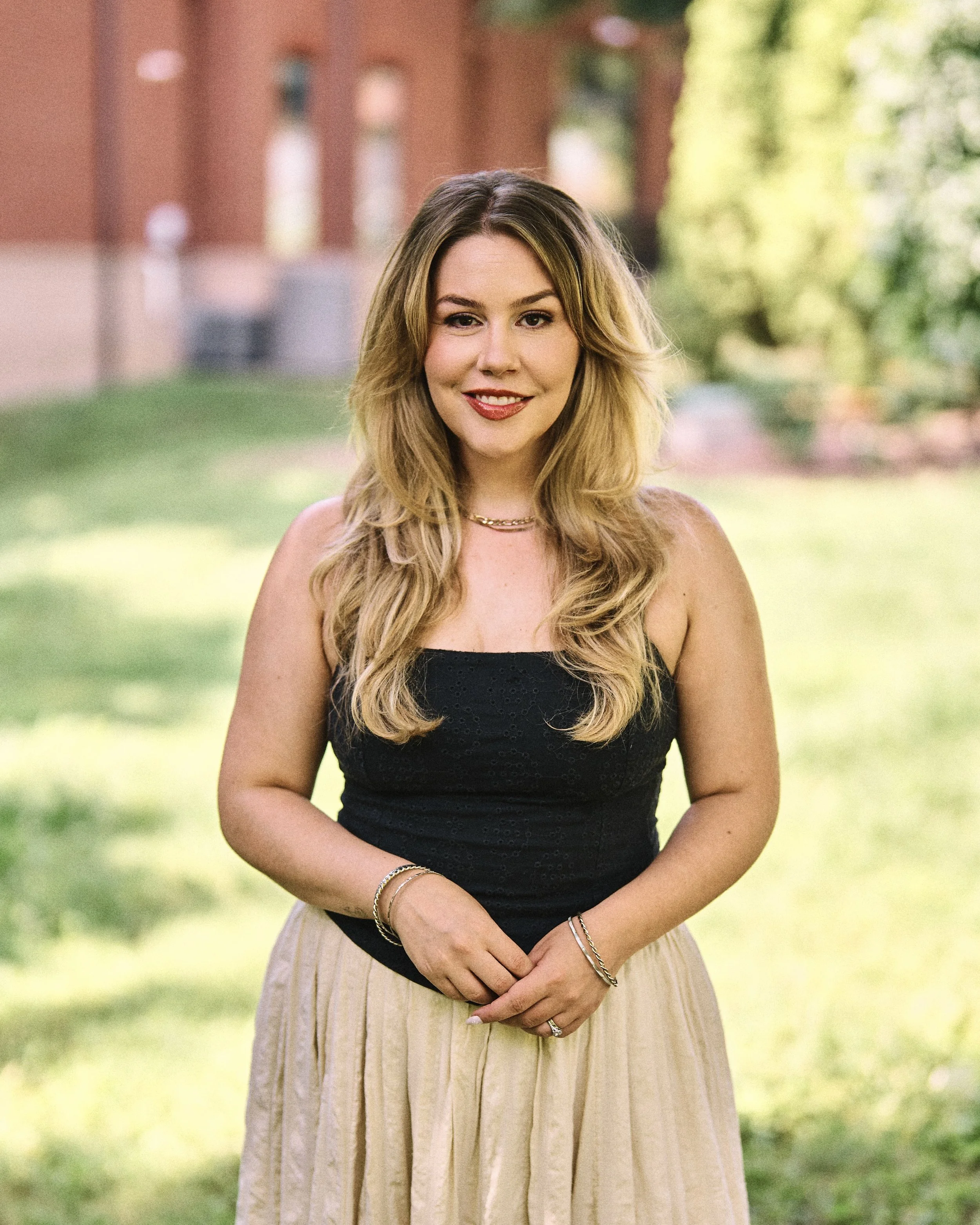 A young woman with long, wavy blonde hair smiling outdoors, wearing a black top, beige skirt, and jewelry, with a blurred background of green trees and a brick building.