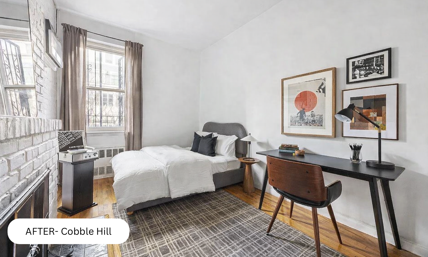 A renovated bedroom with white walls, wooden floors, a gray bed with white and black pillows, a black desk with a wooden chair, framed artwork on the wall, a window with beige curtains, and a small black side table with a record player.