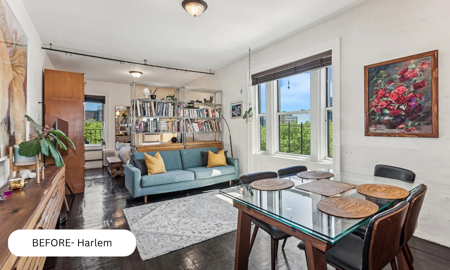 A living room with large windows, a blue sofa with yellow pillows, a dining table with six black chairs and woven placemats, a wooden sideboard, a metal bookshelf with books, and artwork on the walls.