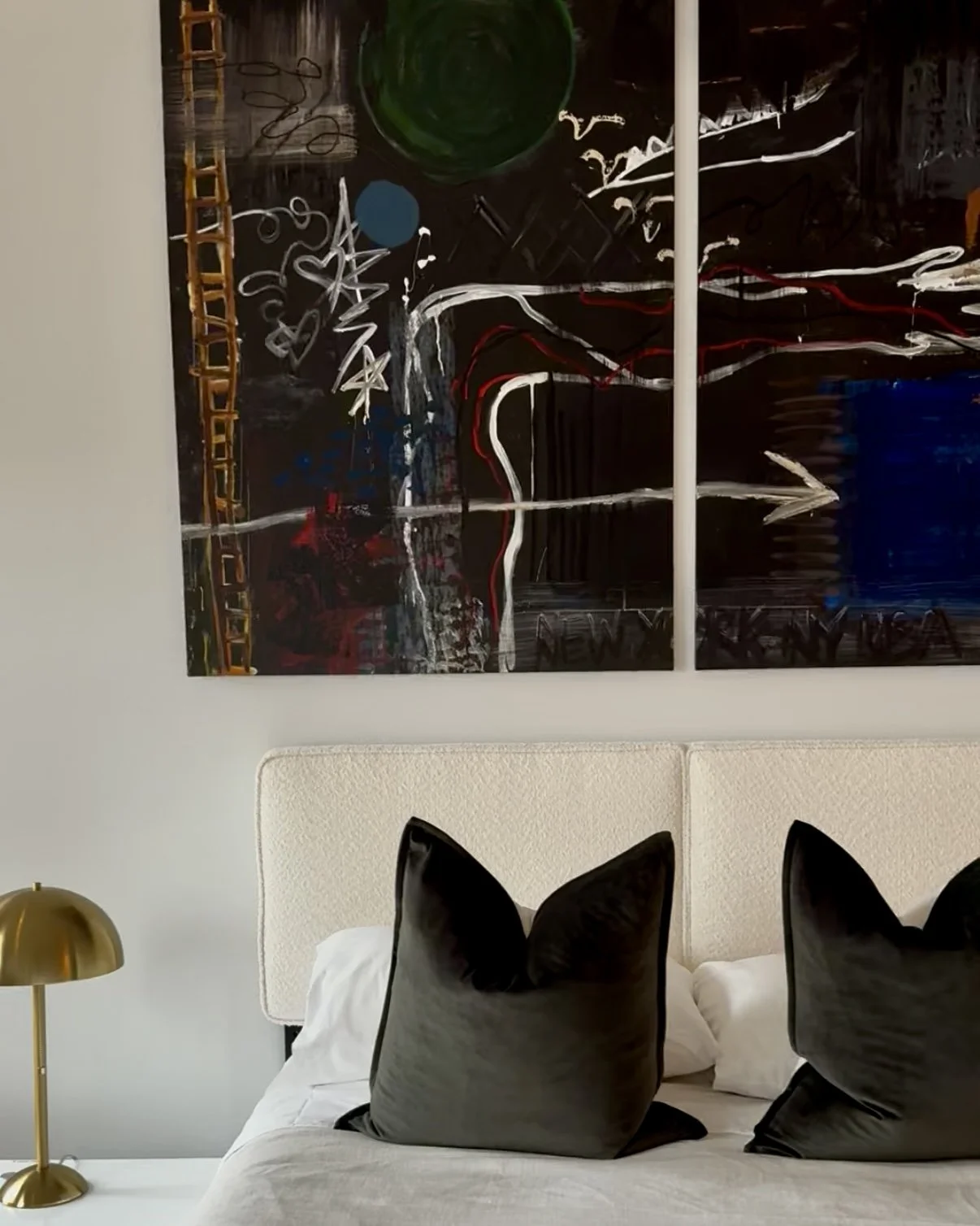 Part of a bedroom with a white bed, black pillows, a gold bedside lamp, and a large abstract painting with dark background and colorful streaks hanging above the bed.