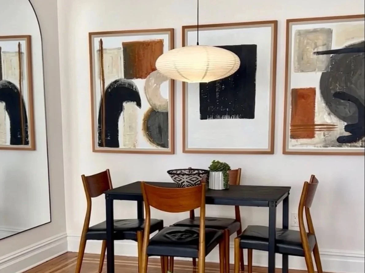 Dining area with black table and four wooden chairs, abstract artwork on white walls, paper lantern ceiling light, mirror, and decorative bowl with plant on table.