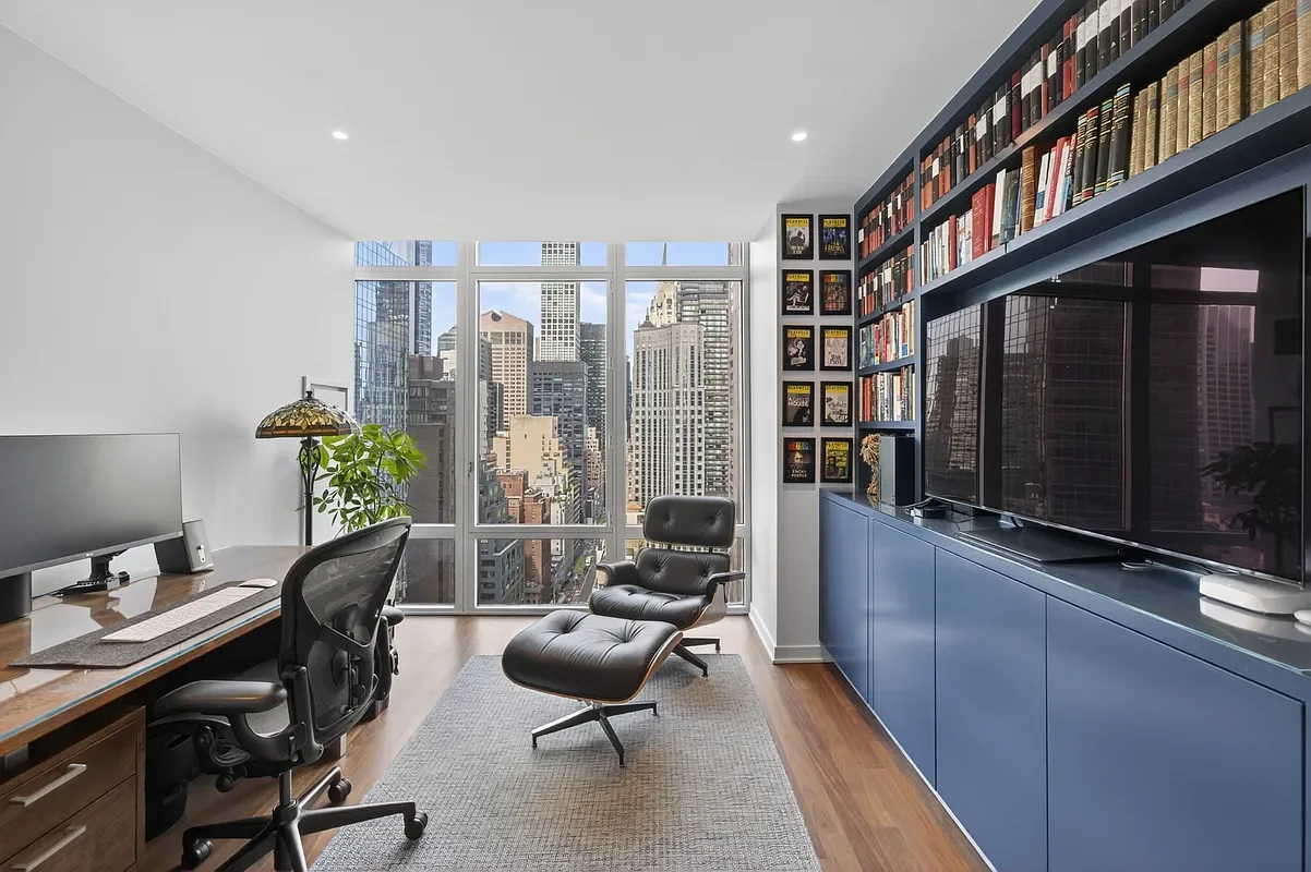 Modern office with large window view of city skyscrapers, a black lounge chair with matching ottoman, a blue storage cabinet with a TV, a bookshelf filled with books, and a workspace with a computer, desk lamp, and plant.