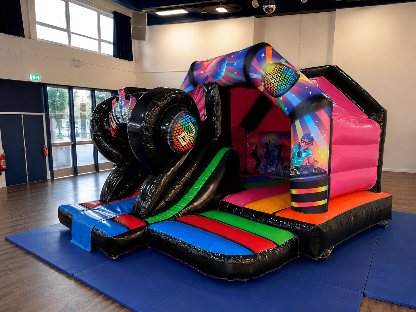 Colorful inflatable bounce house with a DJ cartoon on the side, set up indoors on blue mat, with slide and tunnel.