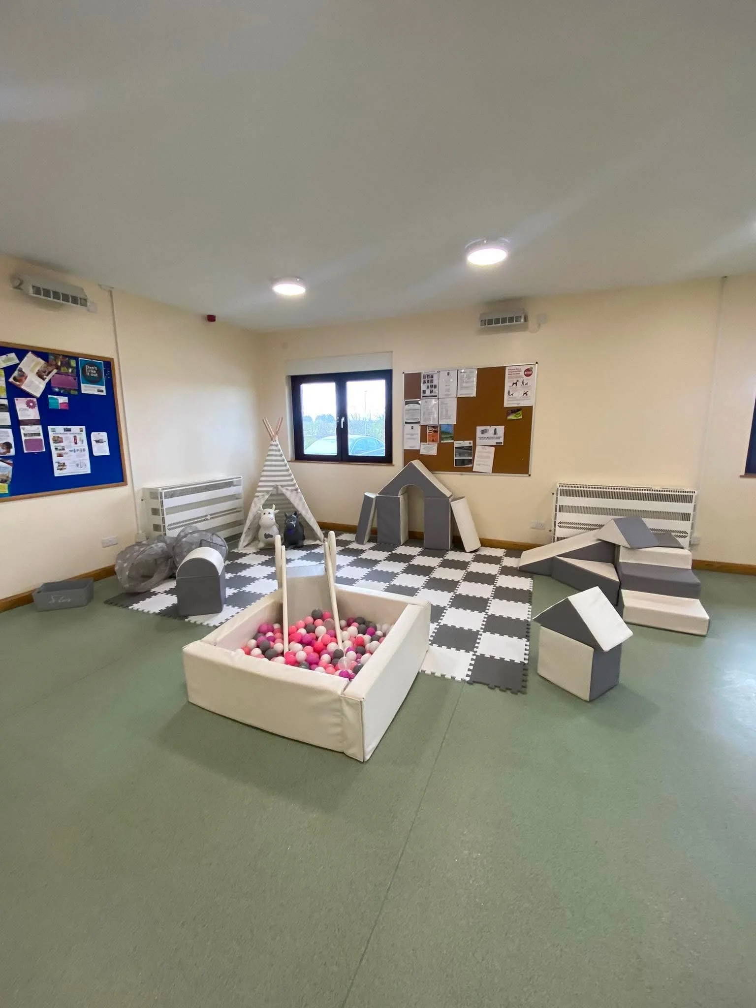 Indoor playroom with black and white checkered rug, soft toy animals, a ball pit, a small wooden house, foam blocks, and bulletin boards on beige walls.