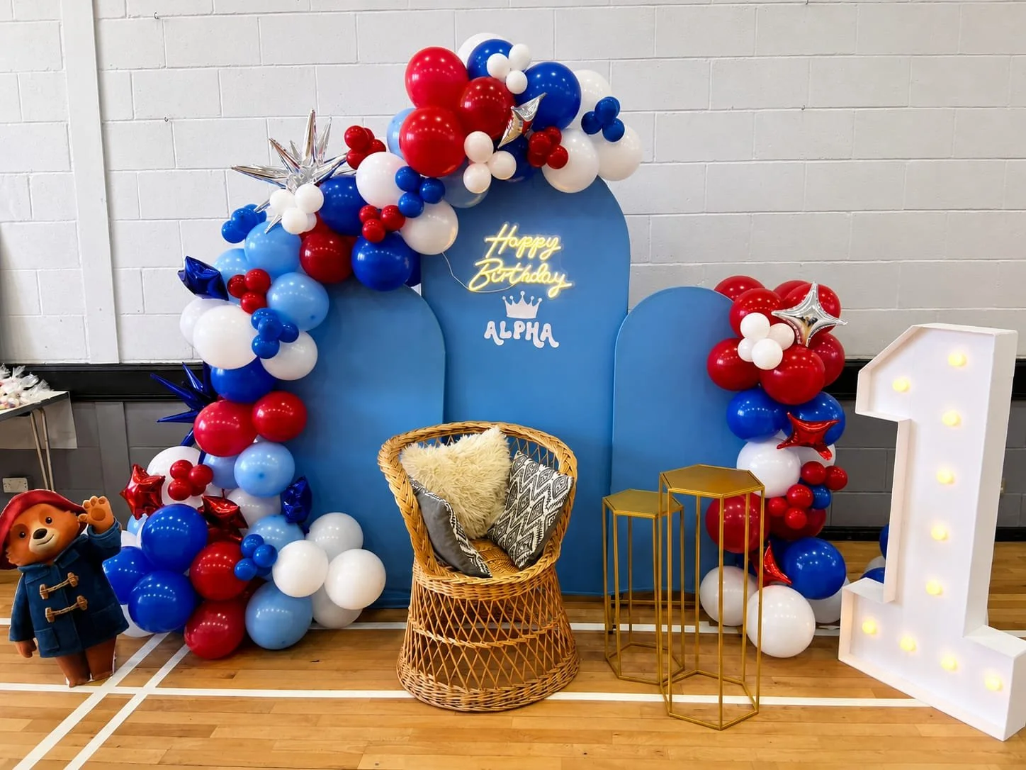 Colorful birthday decoration setup with balloons in red, white, and blue, a blue backdrop with a 'Happy Birthday' neon sign and the name 'Alpha', a wicker chair with pillows, a large illuminated number one, and a small stuffed animal character on the left.