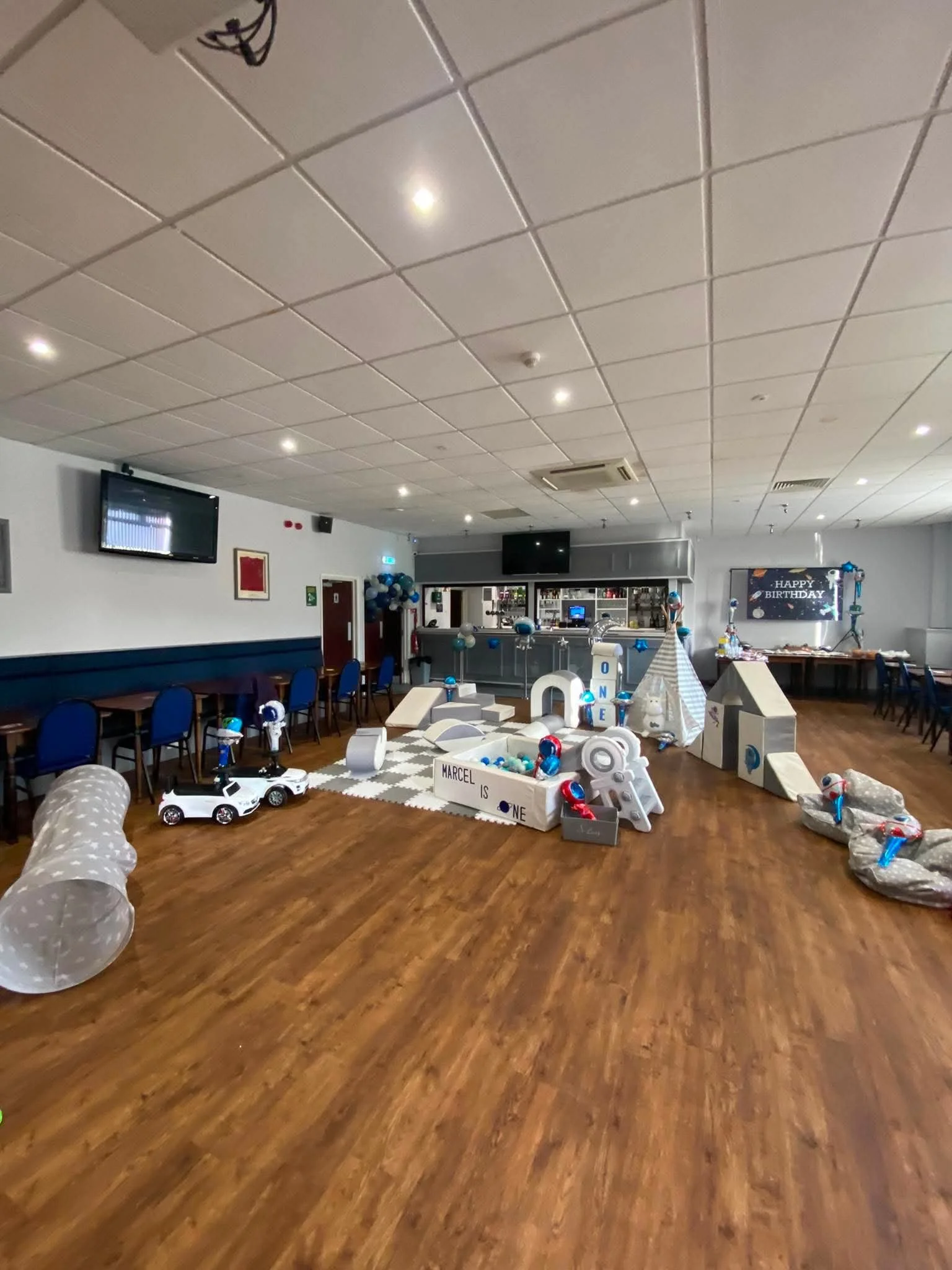 Party room decorated for a birthday celebration with blue, white, and gray balloons, a 'Happy Birthday' banner, and party favors, with a play area including toy cars and large wrapped presents, and a bar area in the background.