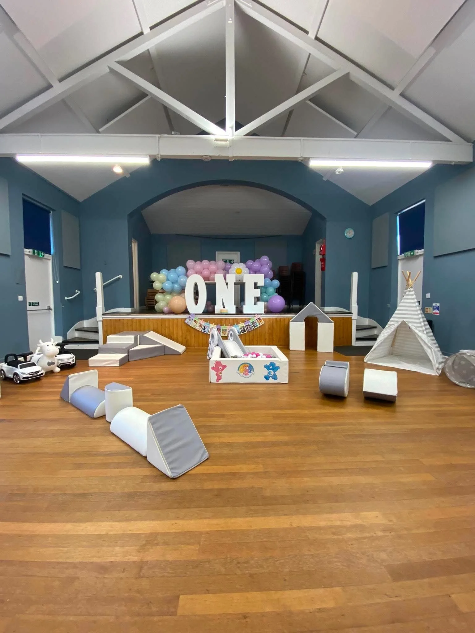 An indoor space decorated for a celebration, with pastel-colored balloons and large white letters spelling 'ONE' on the stage. There are children's toys and play items scattered on the wooden floor, including toy cars, a plush unicorn, and a small pl