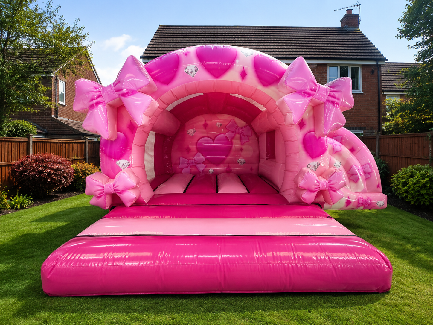 A large pink inflatable bounce house with heart and bow decorations, set up on a grassy backyard with houses and trees in the background.