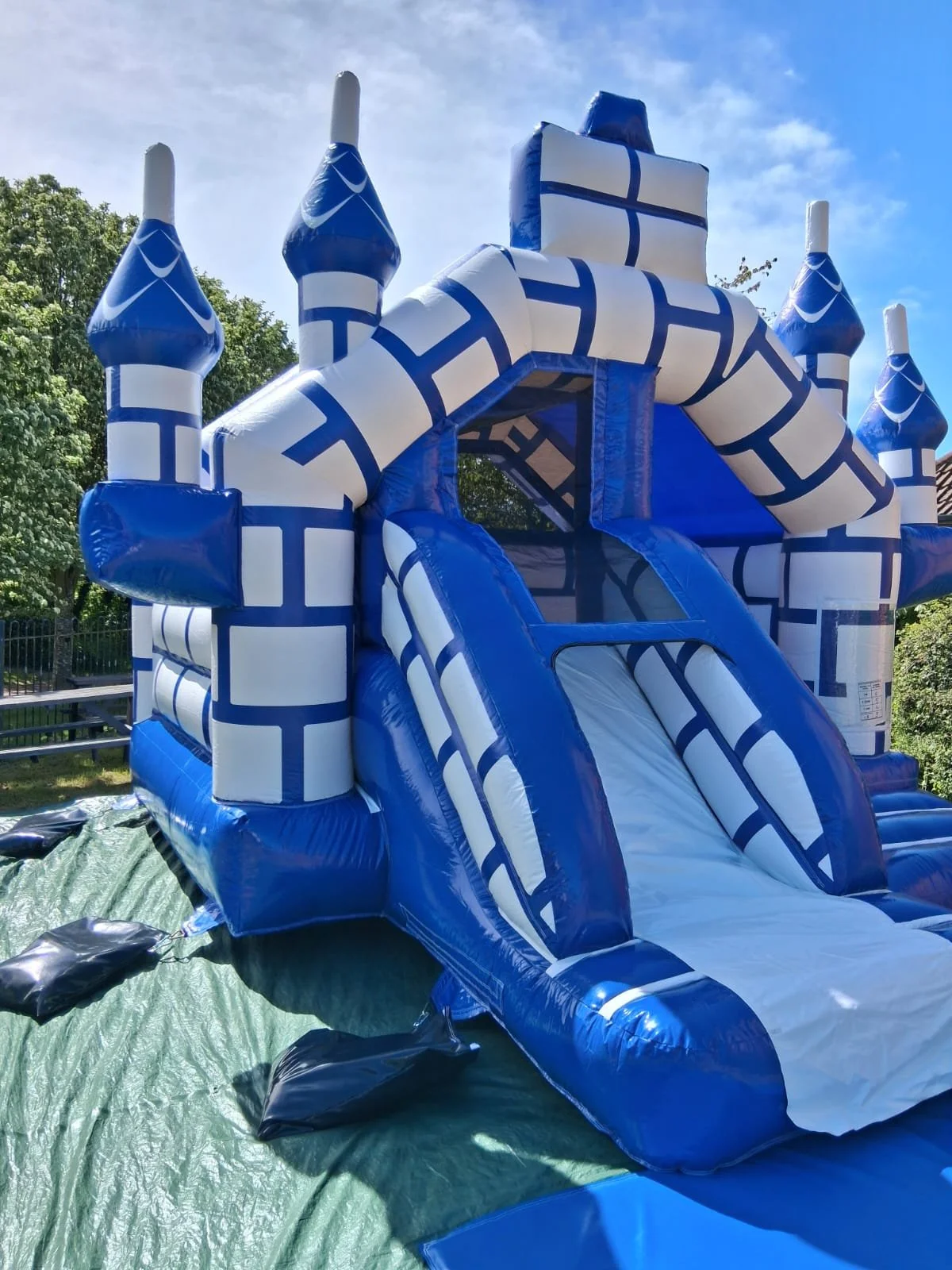 Blue and white inflatable castle with a slide, set outdoors on a green tarp, with trees and a fence in the background.