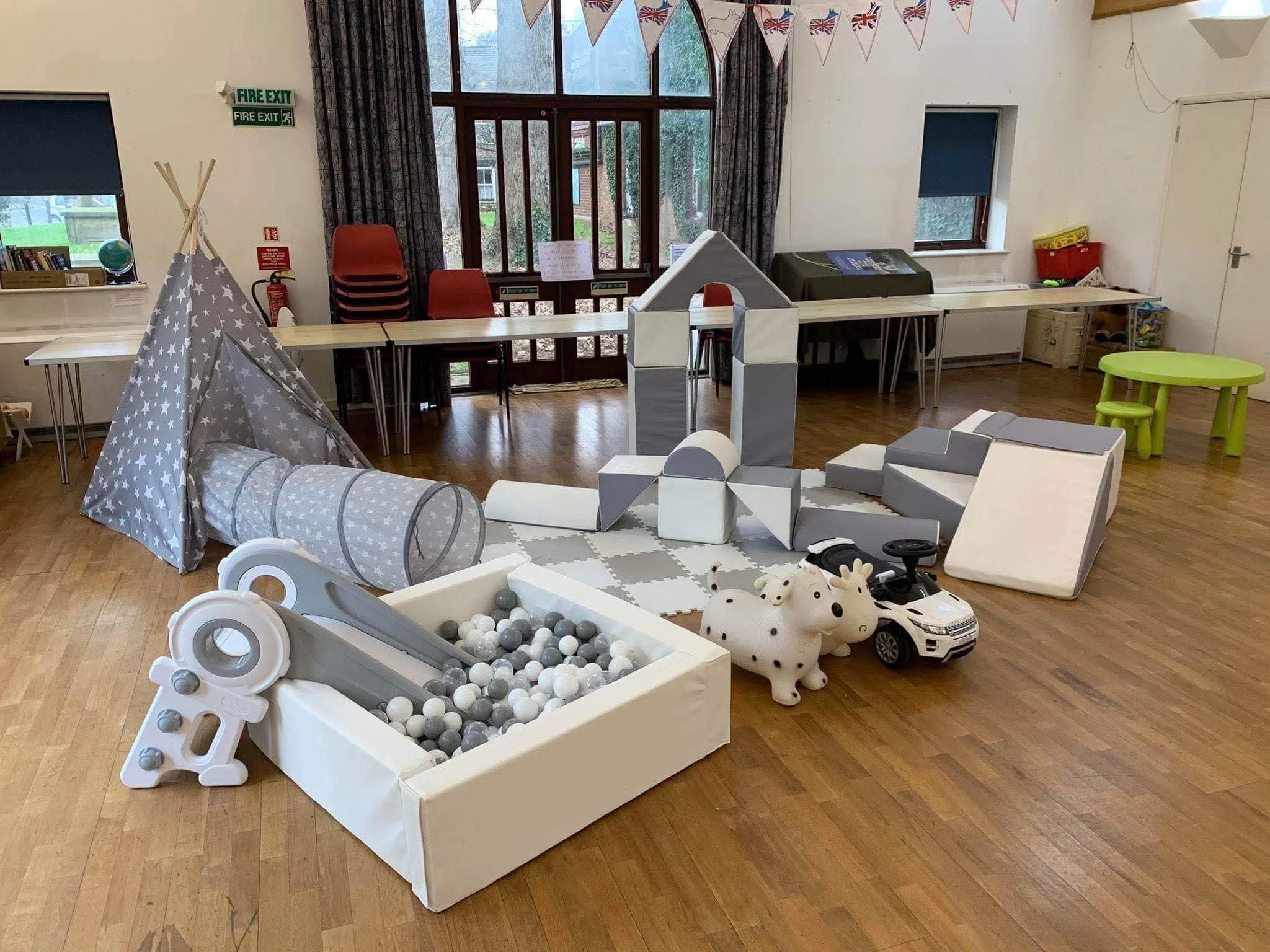 Indoor play area with a gray star pattern tent, a ball pit with white and gray balls, and various toys including plush animals and a toy car. There are stacked red chairs, large windows, and bunting flags hanging from the ceiling.