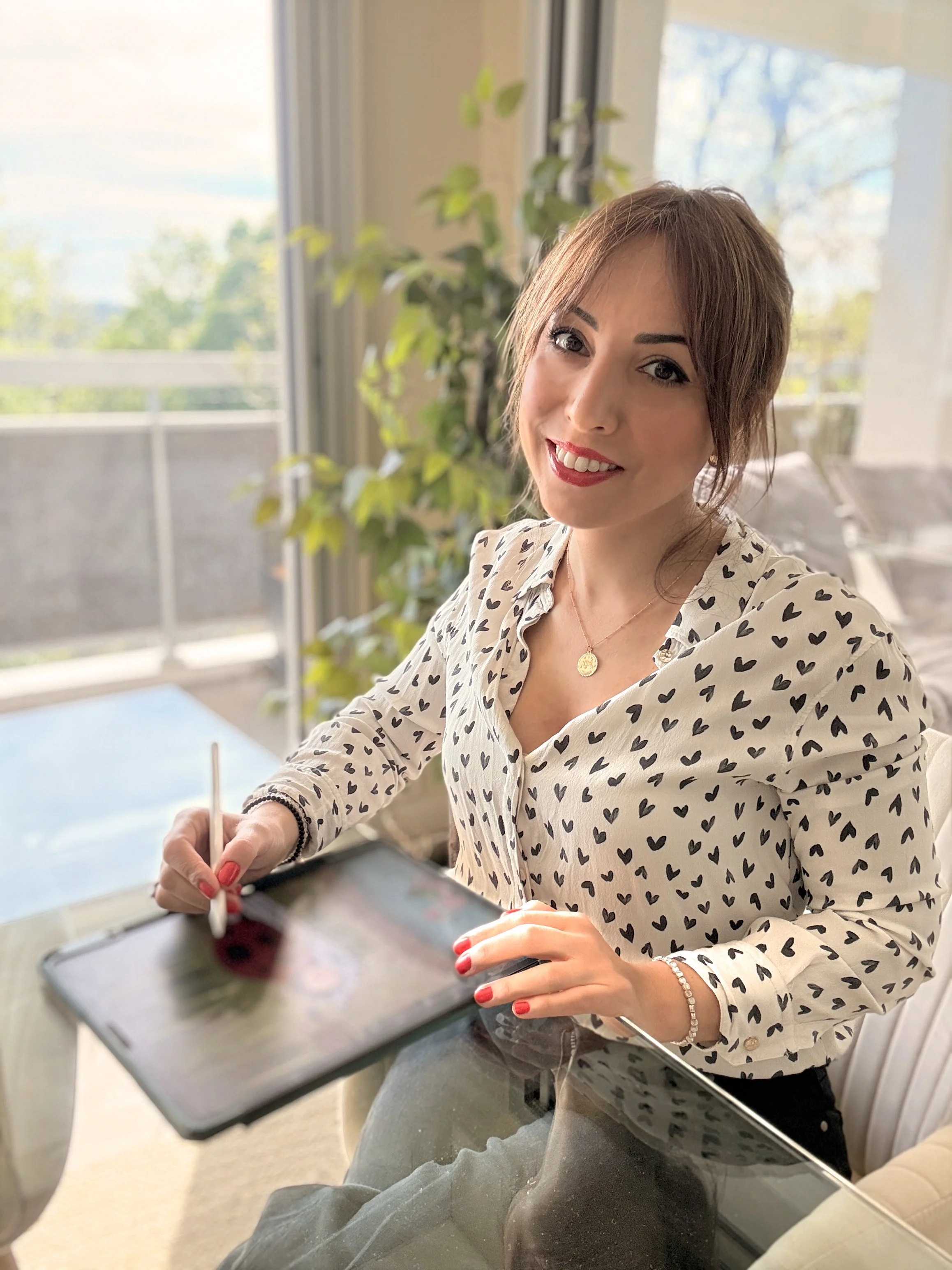Palma Mena, illustratrice, indossa una camicia bianca con motivo a cuori neri e un ciondolo d'oro. Sta utilizzando il suo ipad pro, per le sue illustrazioni digitali. É seduta a un tavolo di vetro, vicino a una finestra.