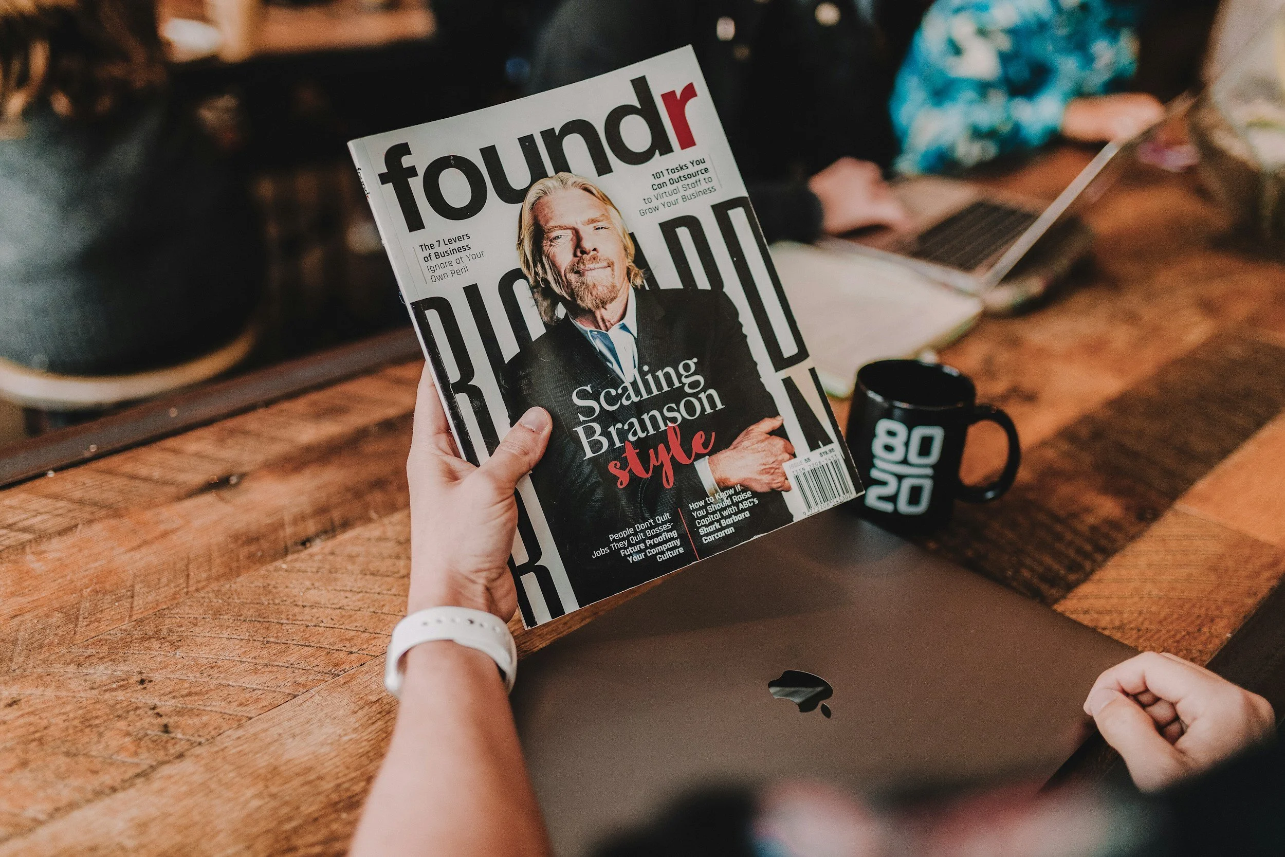 A person holding a magazine titled 'Foundr' in a café or workspace setting, with a laptop and a black coffee mug on a wooden table nearby.