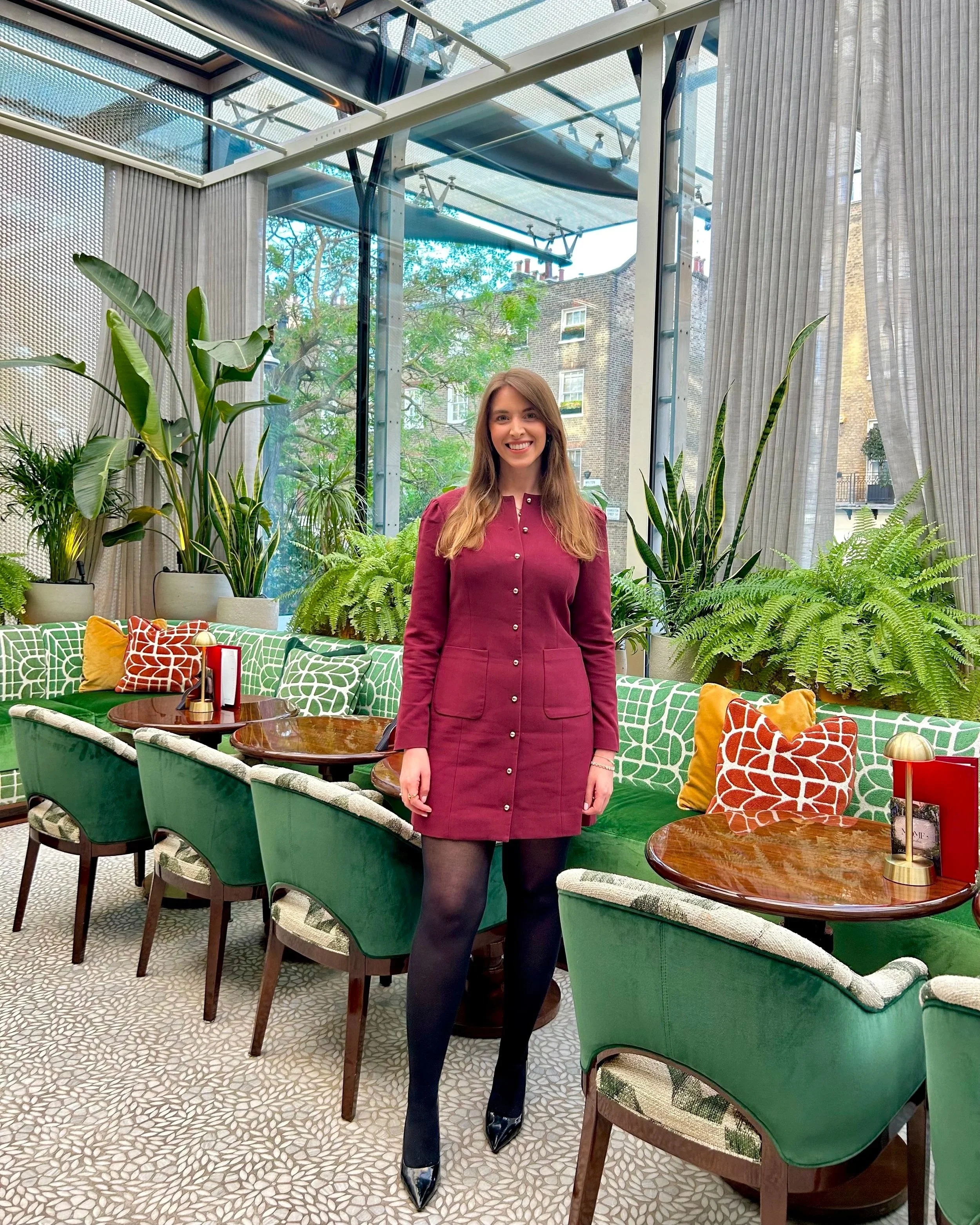 A woman with long brown hair smiling, wearing a burgundy dress, black tights, and black heels, standing in a bright room with large windows, green plants, and patterned furniture.