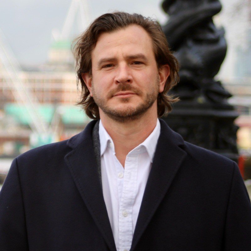 A man with shoulder-length brown hair and a beard dressed in a white shirt and dark blazer, standing outdoors with blurred background of a Ferris wheel and cityscape.