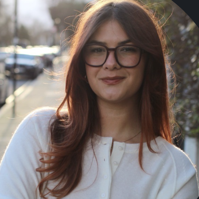 A young woman with long red hair and glasses smiling outdoors during sunset, wearing a white top.