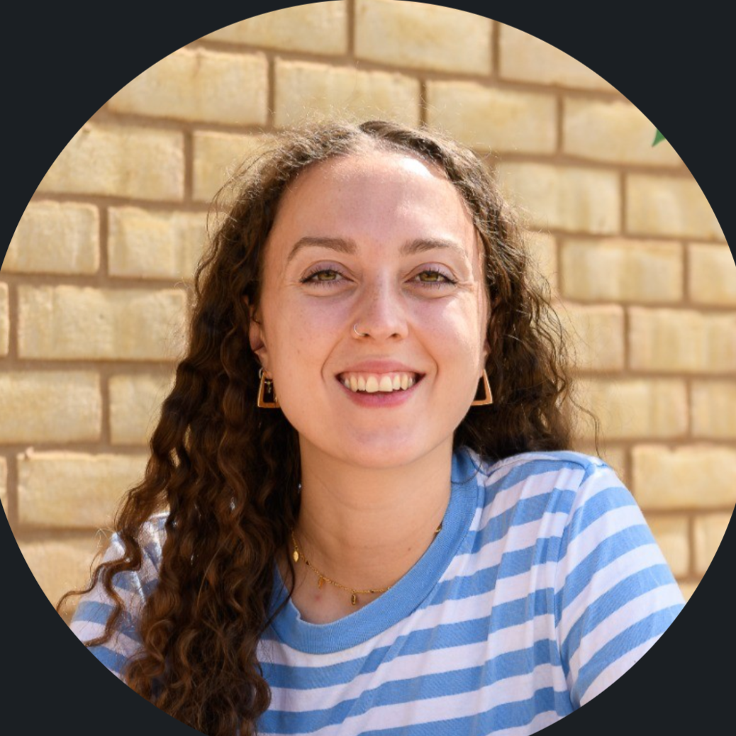 A young woman with curly brown hair and light skin smiling, wearing a blue and white striped shirt, gold jewelry, and earrings, sitting against a brick wall outdoors.