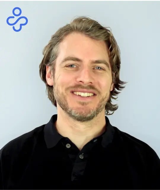 Smiling man with shoulder-length brown hair and a beard, wearing a black collared shirt, standing against a light blue background with a small blue logo in the top left corner.