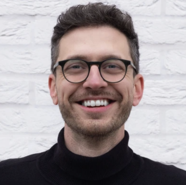 A young man with dark hair, glasses, and a beard, smiling in front of a white brick wall.