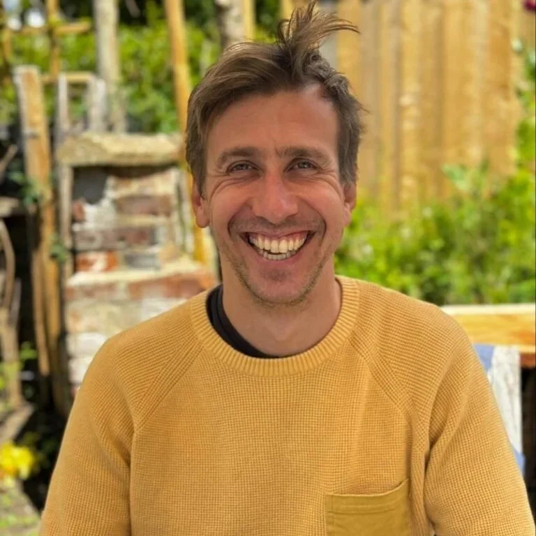 Smiling man with light brown hair wearing a yellow sweater outside in a garden