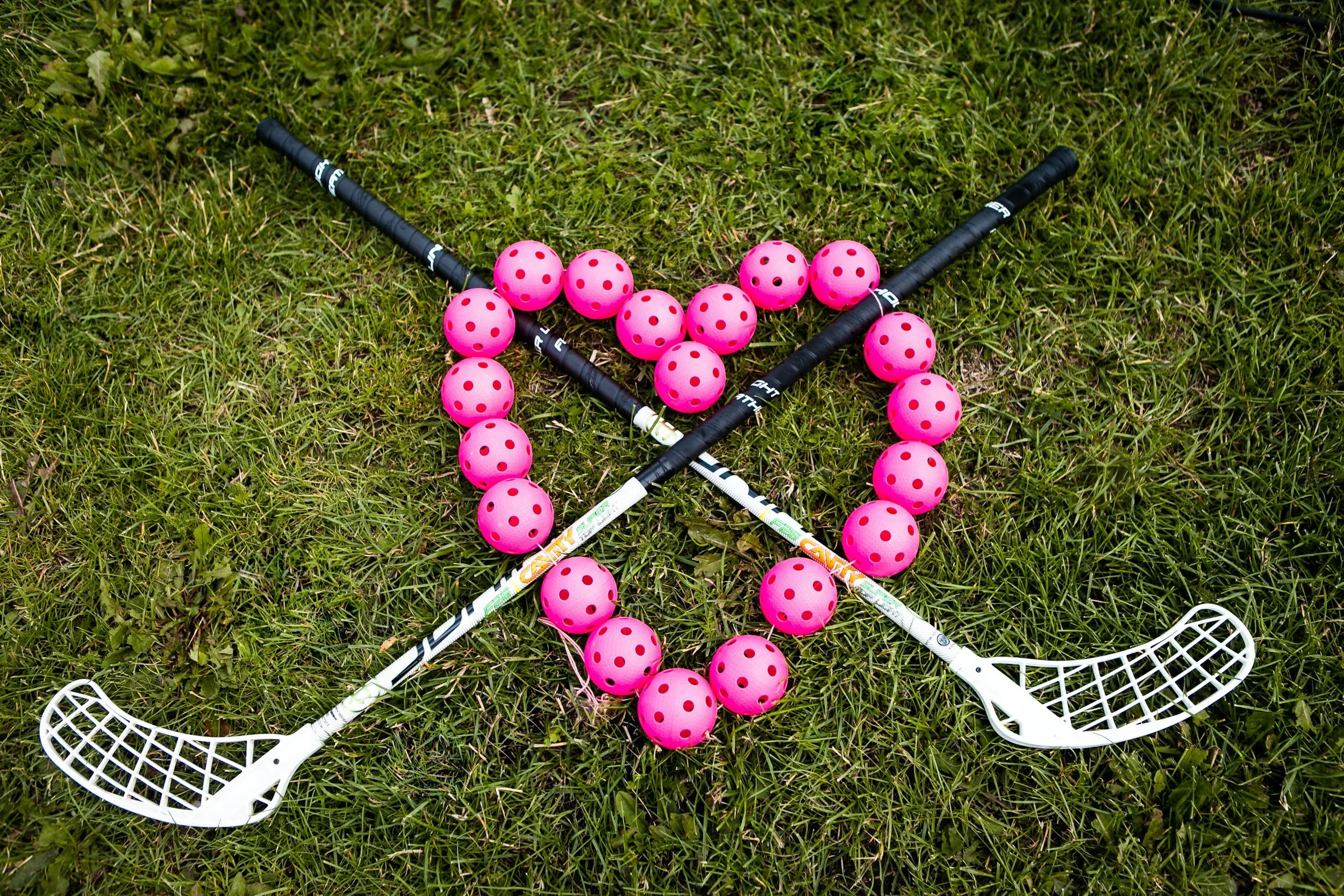 Pink floorball balls with red holes, two white floorball sticks, on green grass.