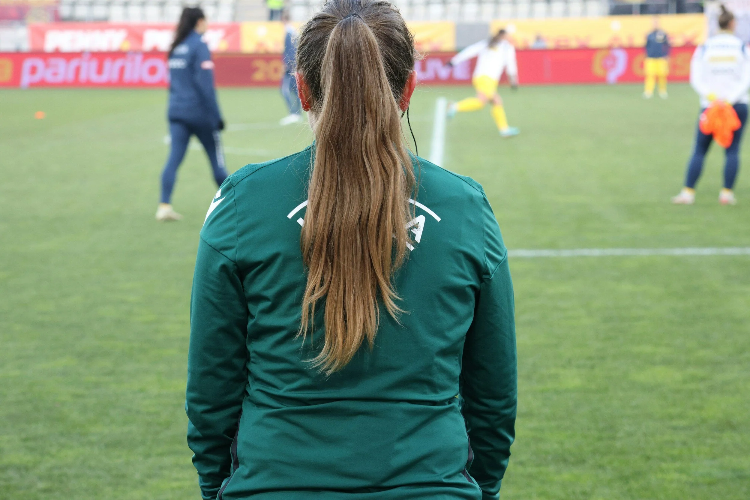 A woman with long brown hair tied in a ponytail, wearing a green sports jacket, stands on a soccer field watching players warm up.