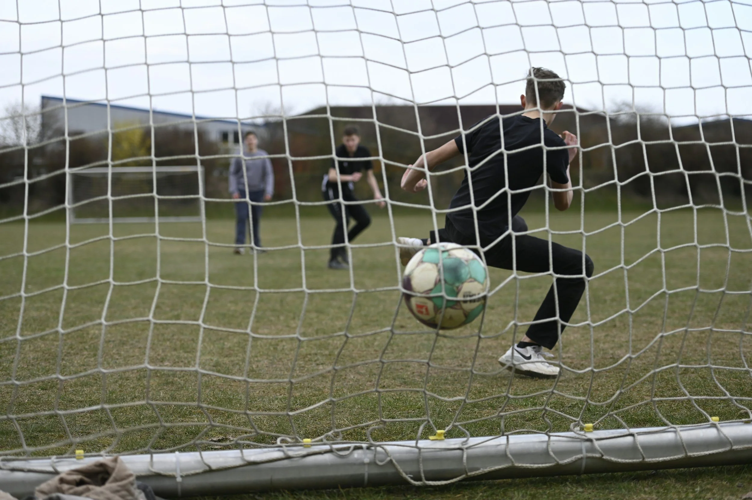 A young boy in black athletic clothing and white sneakers kicks a soccer ball into the goal net during daytime. Two people stand in the background on a grassy field near a soccer goal, watching the game.