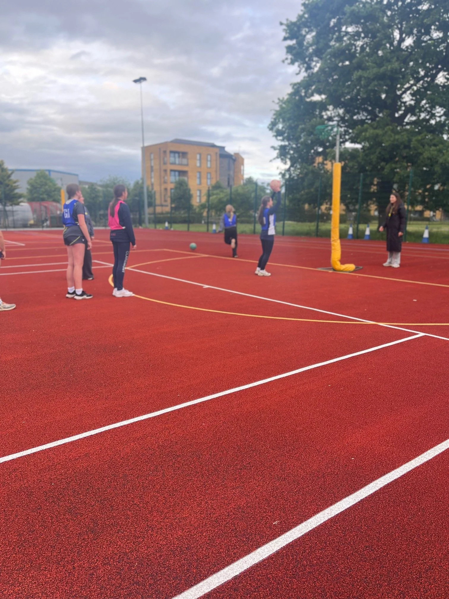 A group of women playing netball on an outdoor court with a red surface, surrounded by trees and buildings. One woman is about to catch or shoot the ball.