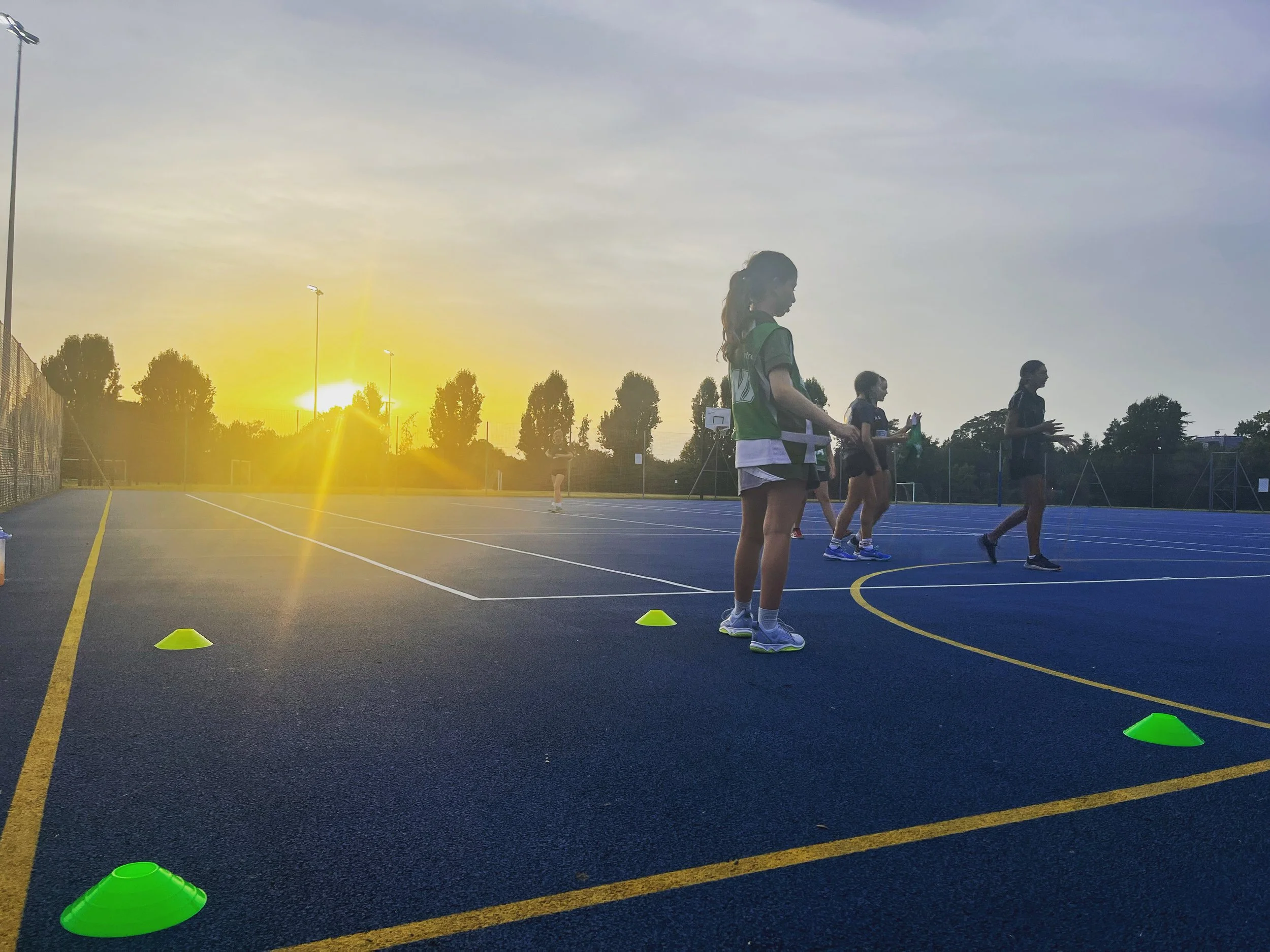 Young women in sportswear participating in an outdoor fitness class on a tennis court at sunset, with orange cones set up for drills.