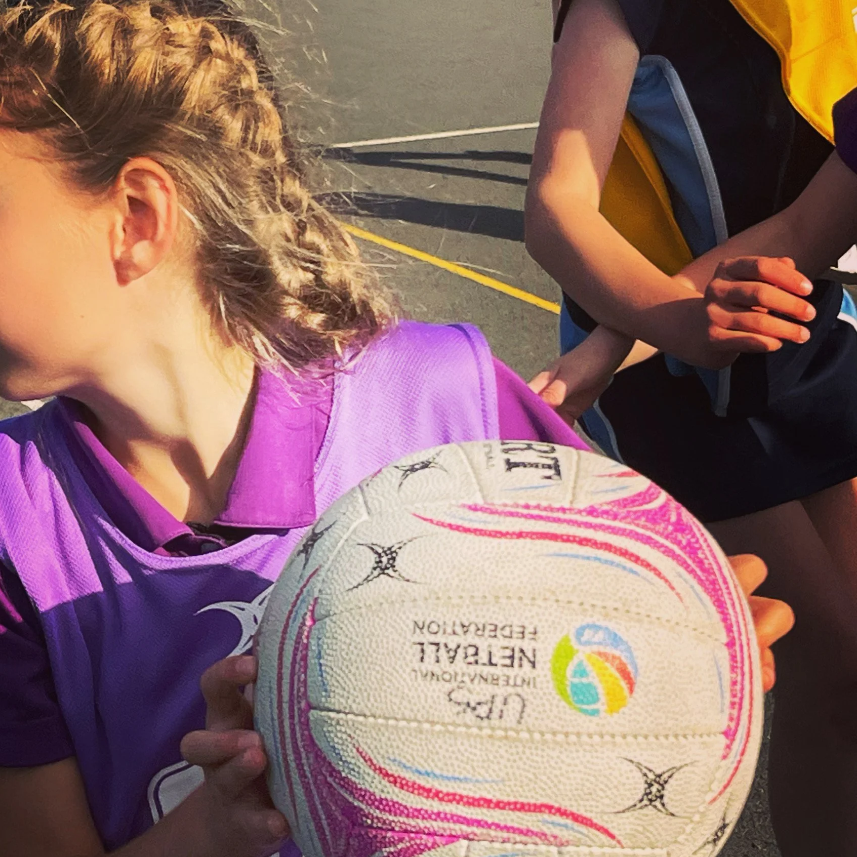 A young girl with braided hair wearing a purple sports jersey holds a volleyball with a colorful logo and the words "Netball World Federation" on it. A boy wearing a black shirt and a yellow backpack is beside her, with his hands clasped. They are on a sports court, possibly during a netball or volleyball event.