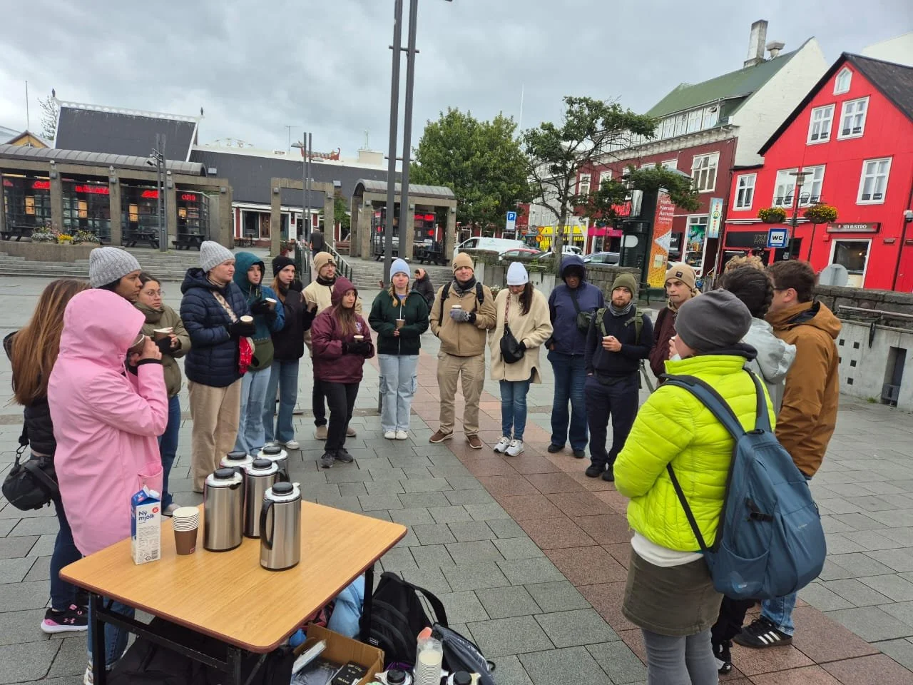A group of people stands in a circle outdoors in a city square, some wearing jackets and hats, with a table of thermoses and cups in the foreground. The background features colorful buildings and a cloudy sky.