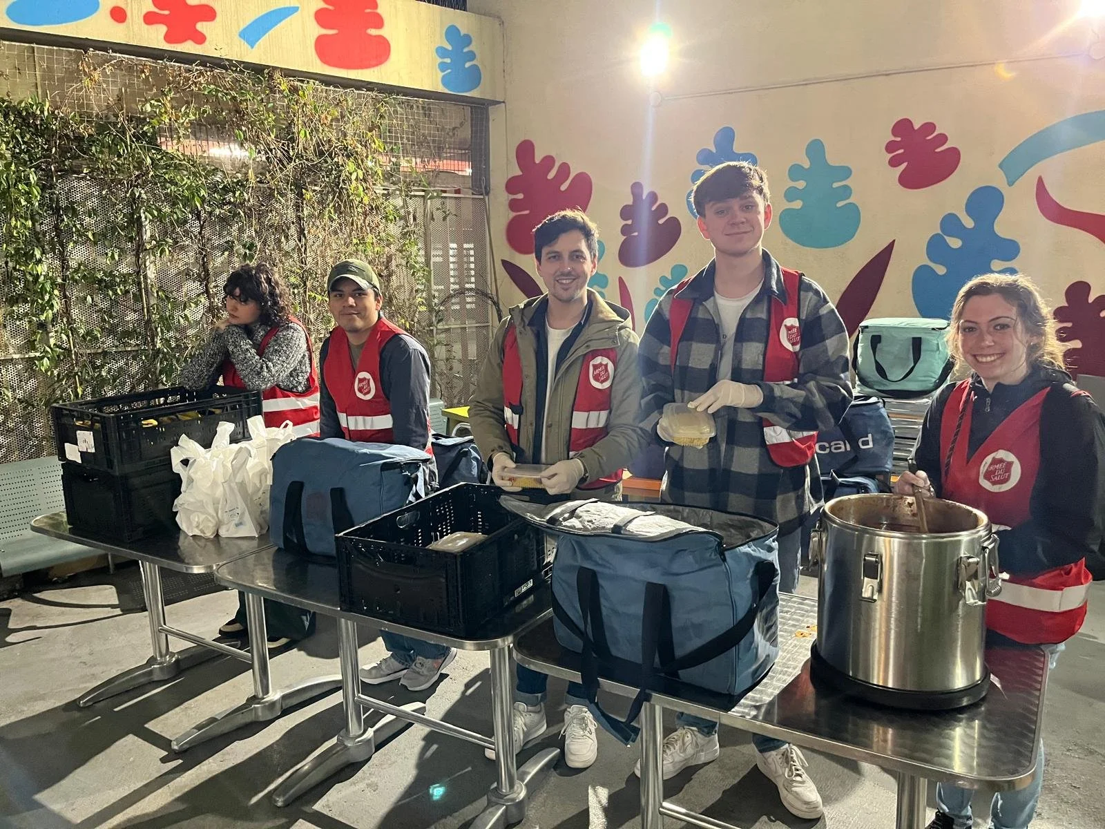 Group of five volunteers wearing red vests and gloves, preparing food outdoors at a table with bags and containers, with a colorful mural background.